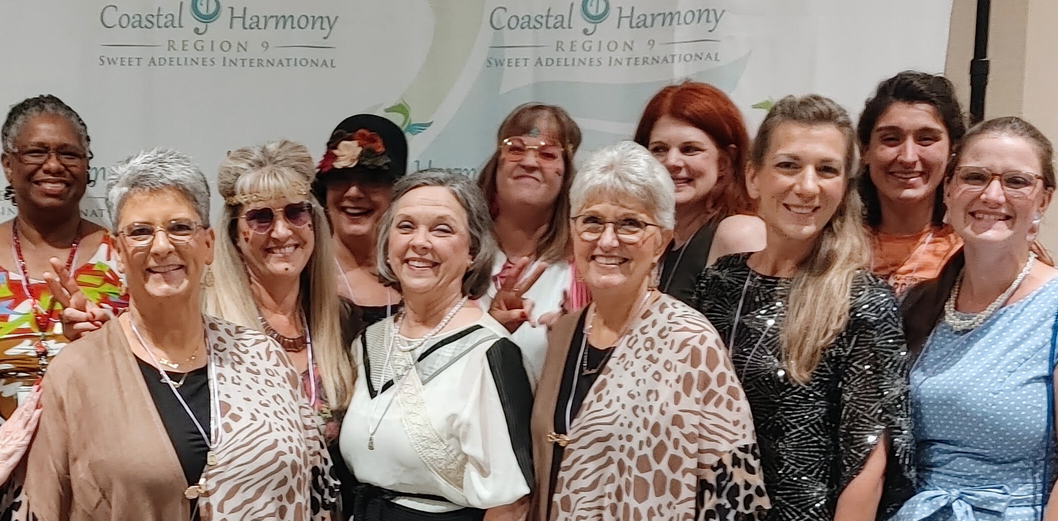 11 women in semi-formal attire stand in a row, smiling at the camera. Behind them, a banner says Coastal Harmony Region 9, Sweet Adelines International