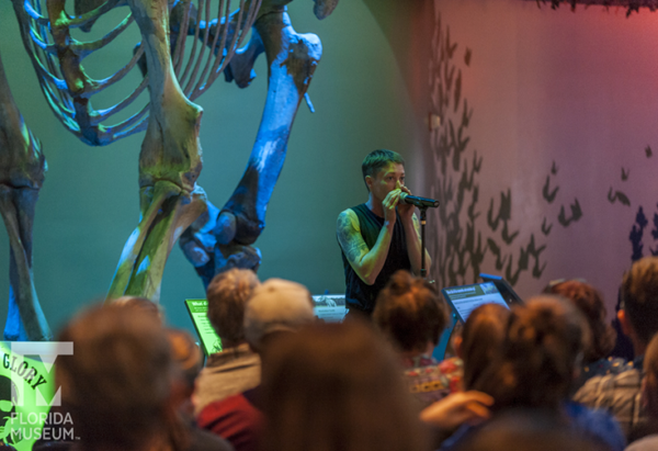 Storyteller at the microphone standing in front of the mammoth skeleton at the museum in front of a crowd