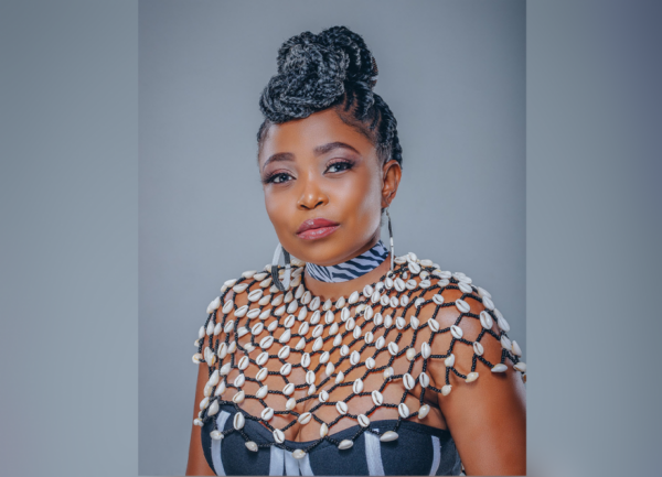 Photo by Emp Cyclone. Medium skinned African woman with dark eyes, and black hair worn in braids styled on top of head. Wearing beaded earrings, zebra striped neckband, white-shell neckpiece, and black top with white stripes.