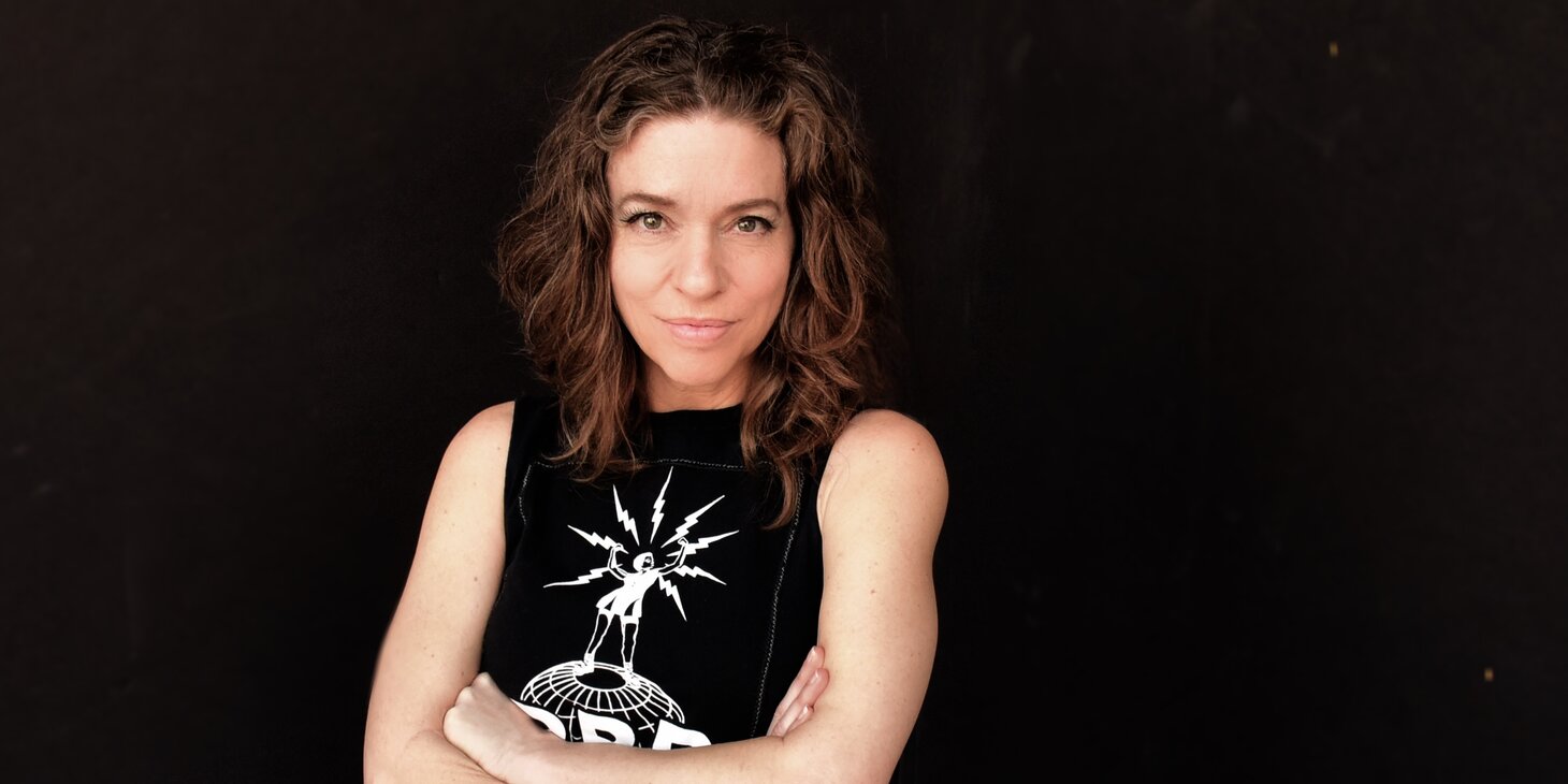 Ani DiFranco standing with her arms crossed looking at the camera with a smirk on her face