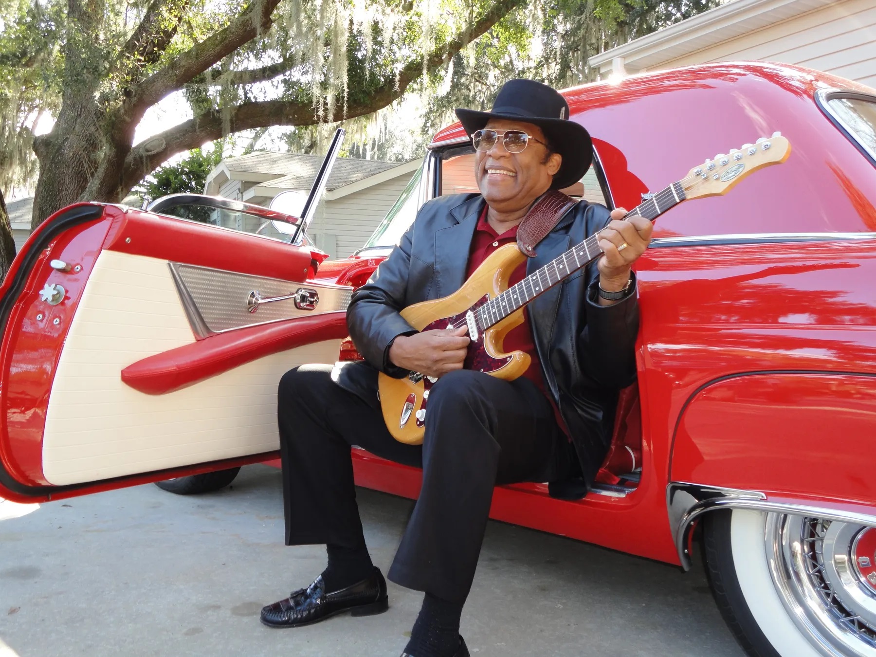 Bobby Blackmon playing a guitar while sitting next to a vintage red car